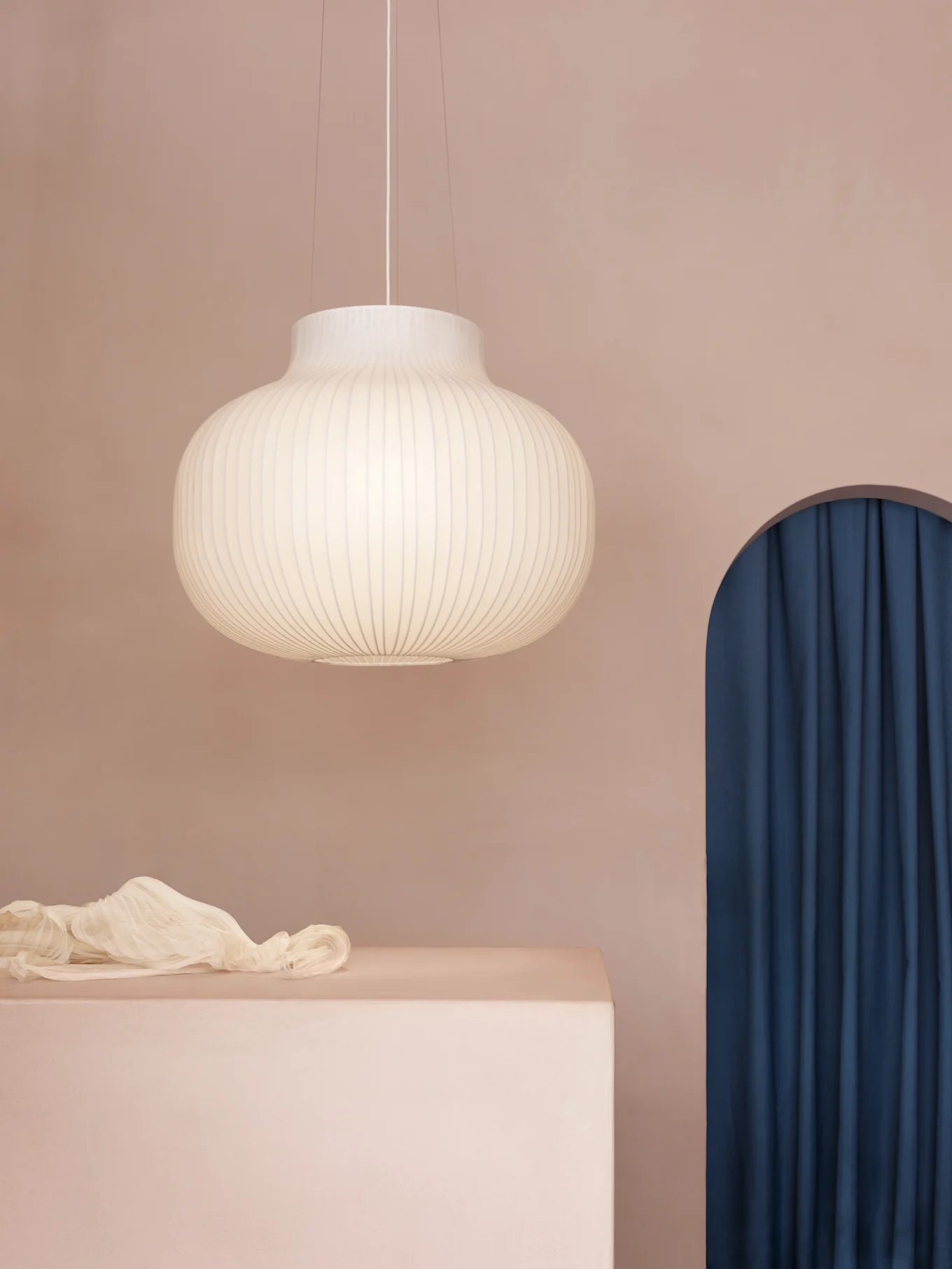 A ribbed, rounded pendant lamp hangs above a pink block with draped sheer fabric, beside an arched blue curtain.