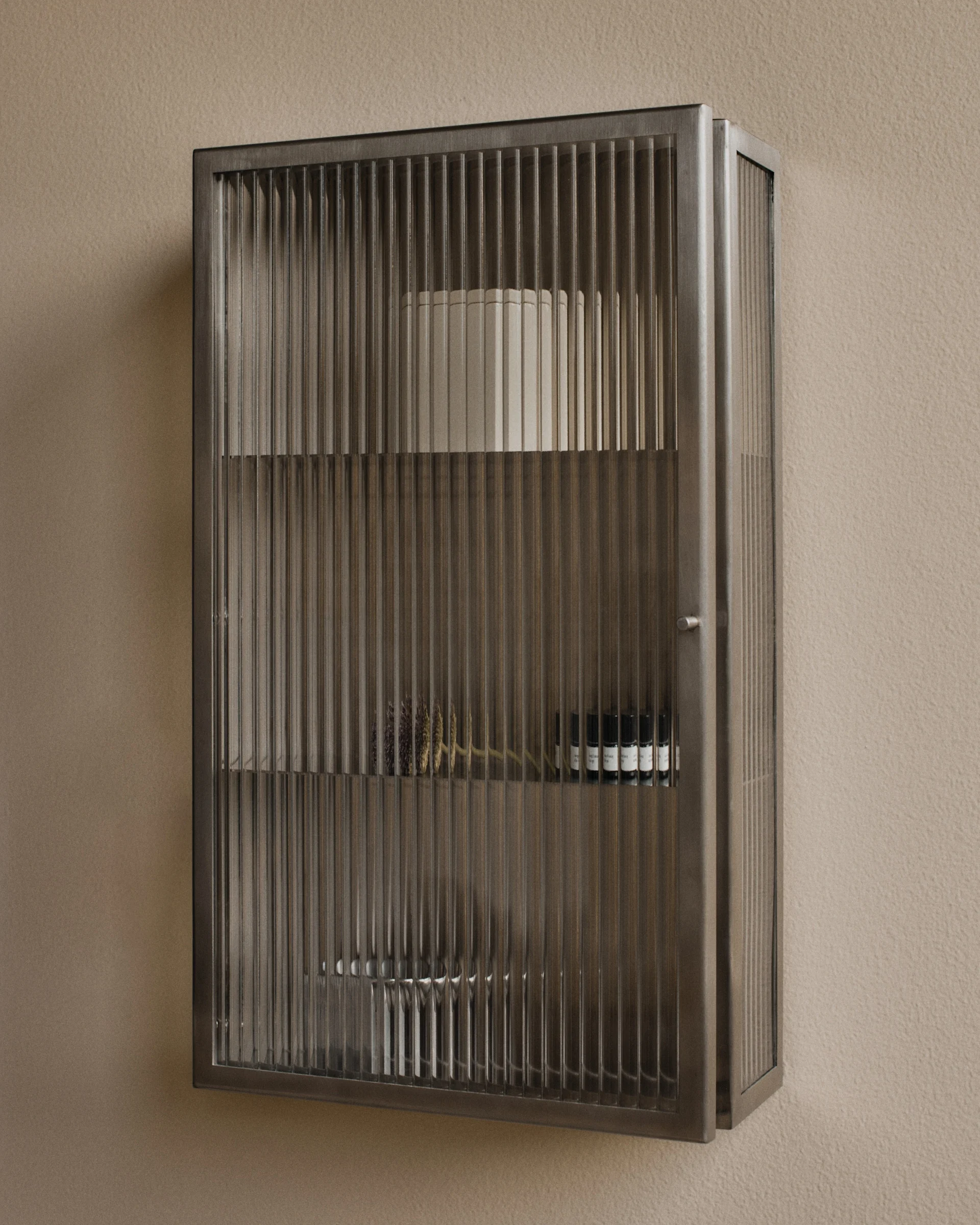 Wall-mounted metal cabinet with fluted glass doors, containing decorative items and small bottles.