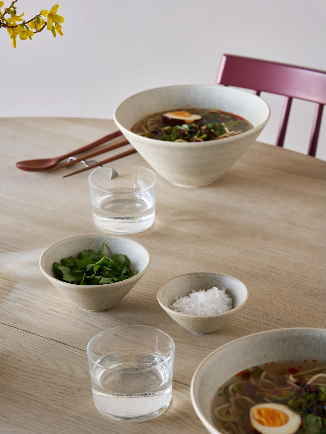 Two bowls of ramen with eggs and herbs, side bowls of cilantro and salt, water, chopsticks, and a spoon on a light wooden table.