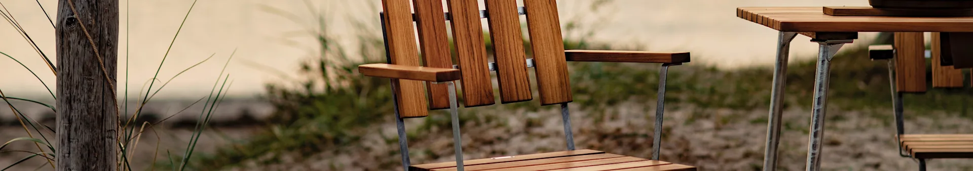 Close-up of a wooden slatted outdoor chair and table on a sandy dune with tall grass and a wooden post.