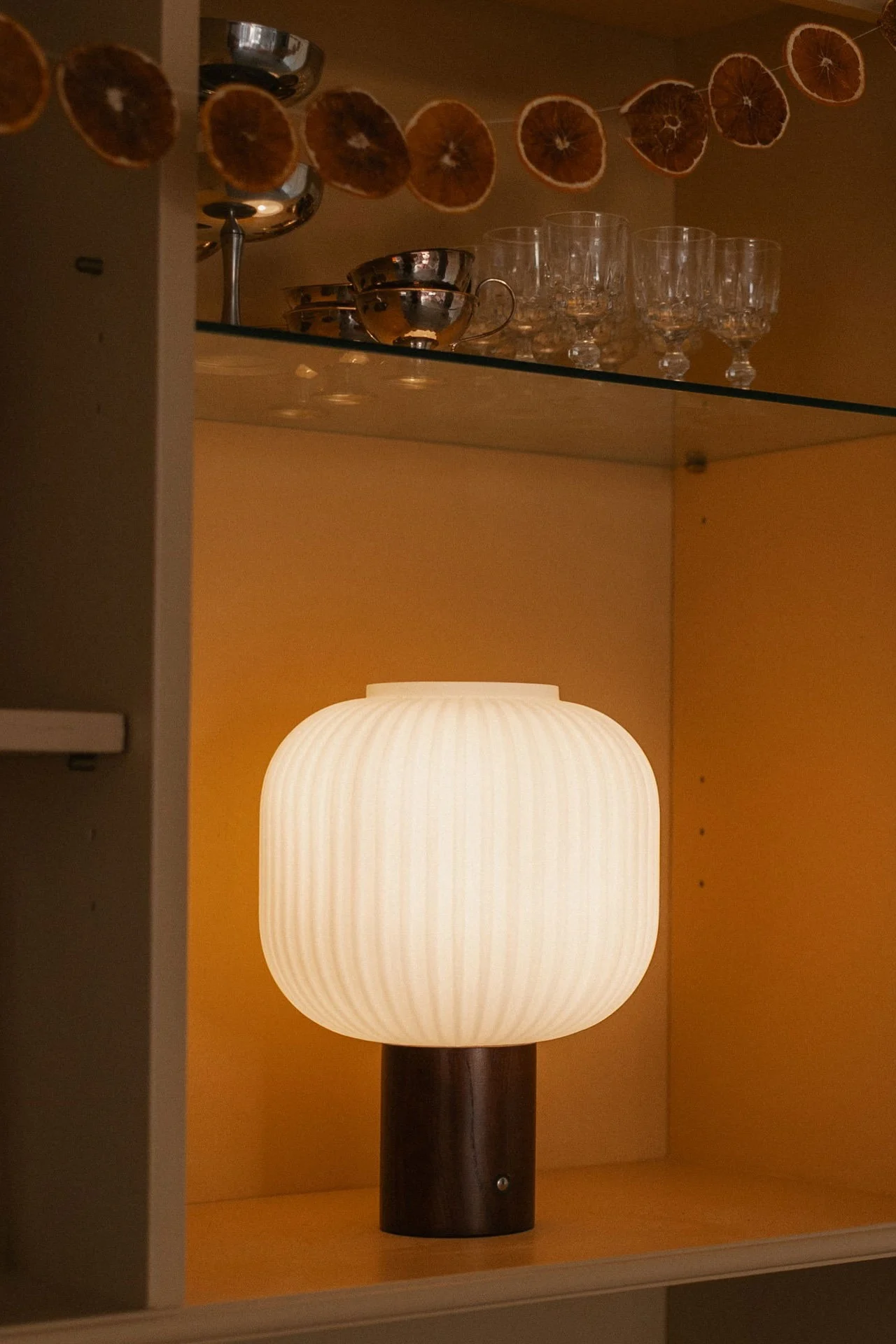 A glowing, ribbed, oblong lamp with a dark wooden base sits in a warm orange cabinet, decorated with dried orange slices.