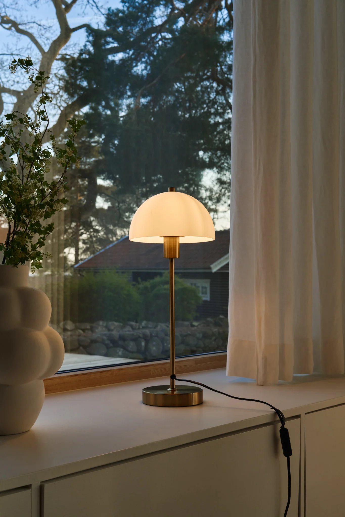 A brass table lamp with a lit white domed shade sits on a windowsill next to a white vase with branches, with an outdoor view.