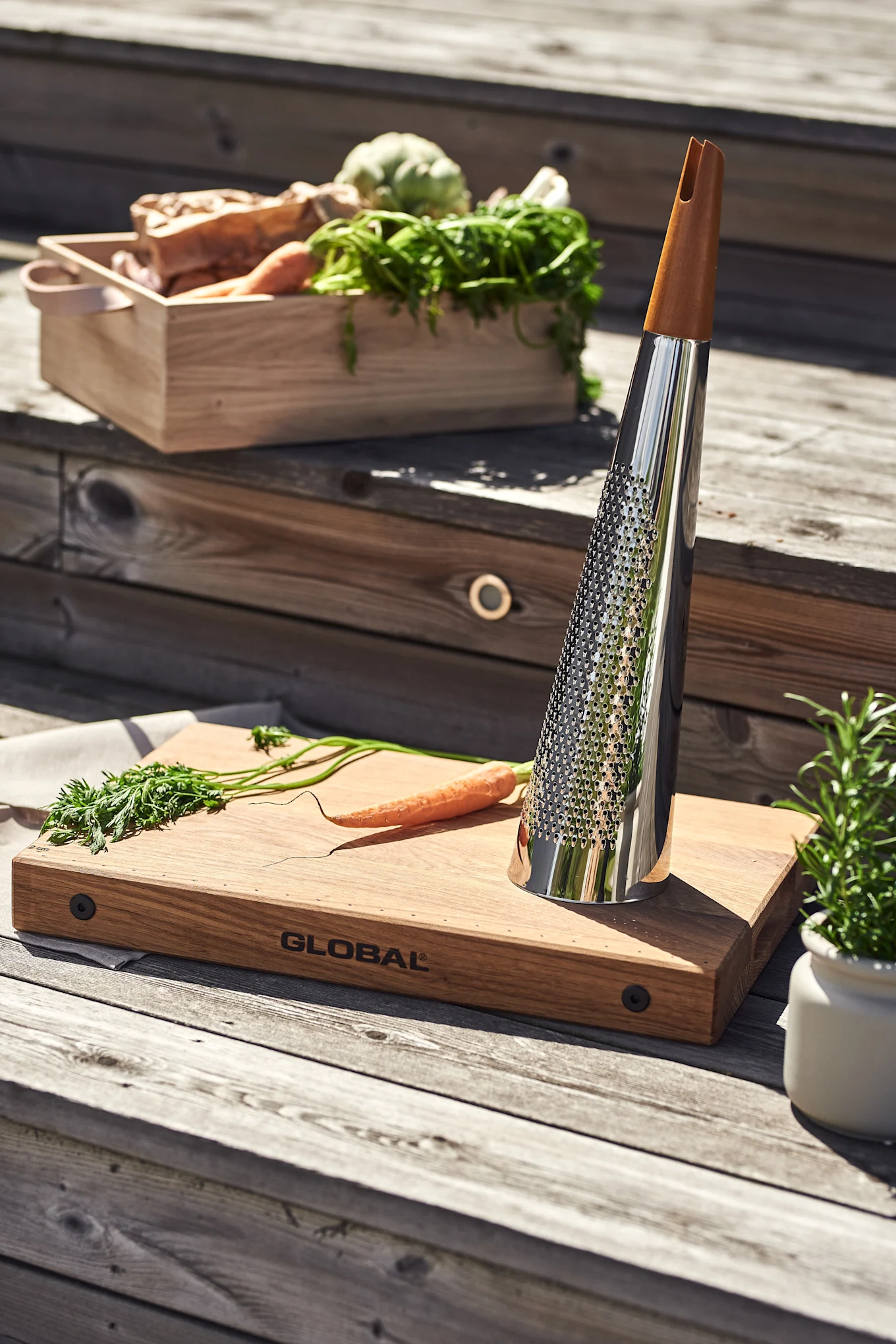 A sleek chrome grater with a wooden handle sits on a wooden cutting board with a carrot. A crate of fresh vegetables is on steps.