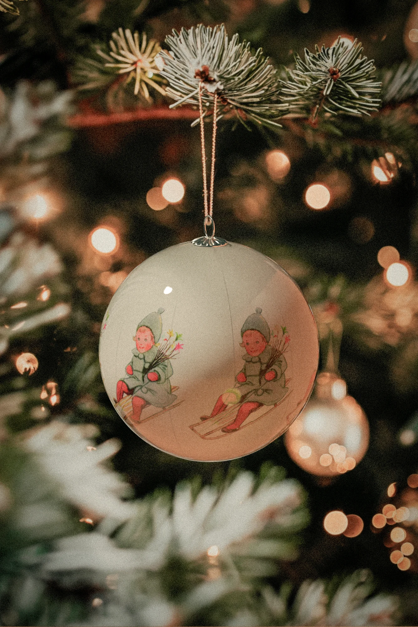 A close-up of a round Christmas ornament depicting two children in winter clothes on sleds, hanging on a blurred Christmas tree with warm lights.