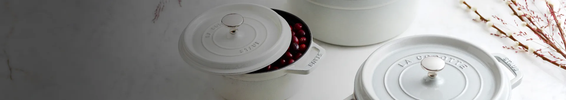 Three white STAUB cast iron cocottes on a marble surface, one open with red berries, another partially visible, and decorative branches.
