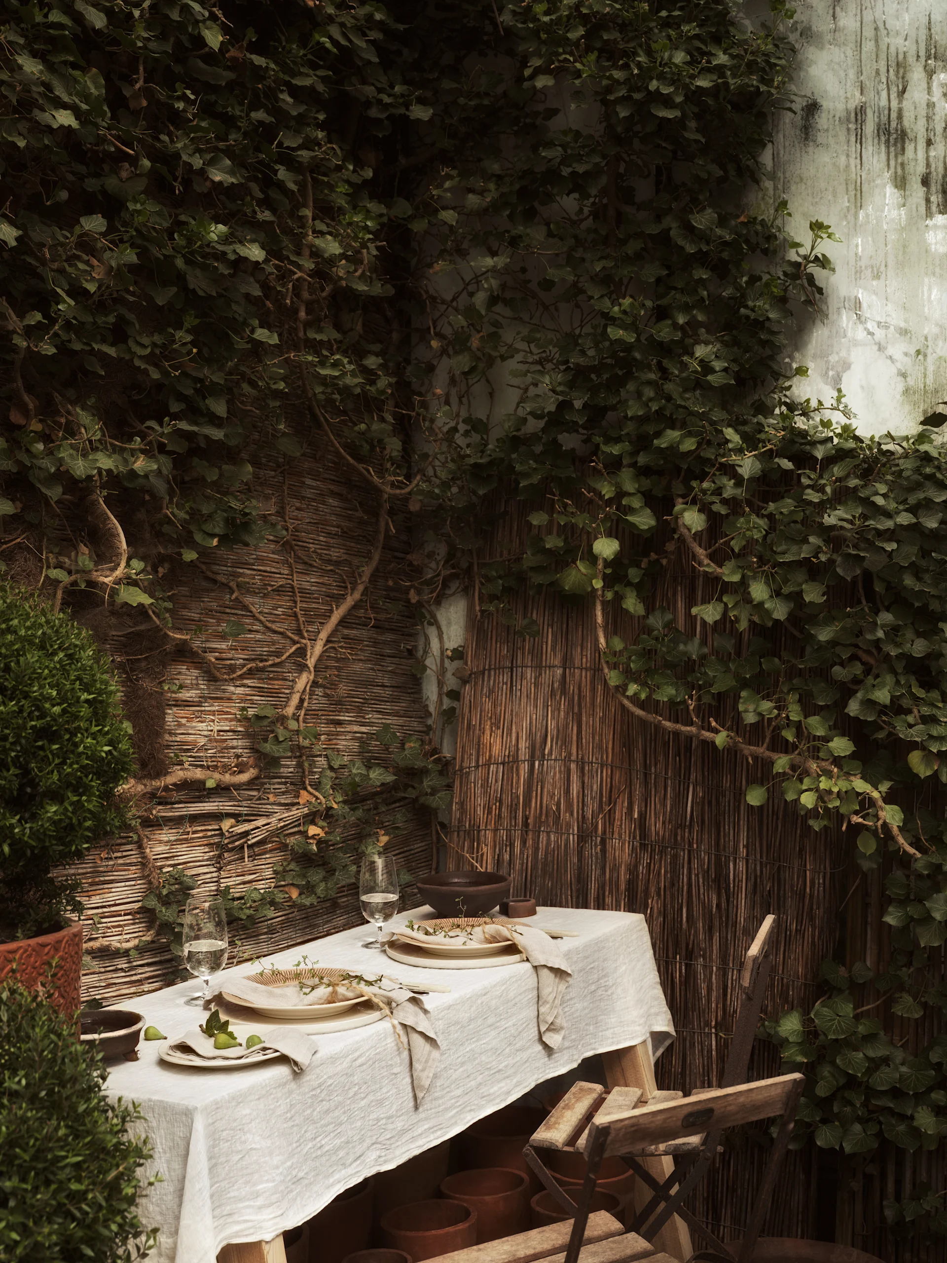 Outdoor dining table set for two with white tablecloth, plates, glasses, and natural decor against ivy and bamboo.