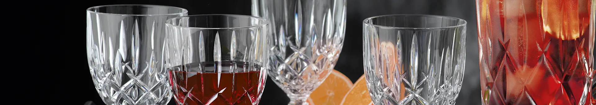 Close-up of three ornate crystal glasses, one containing red liquid, with orange slices in the background.