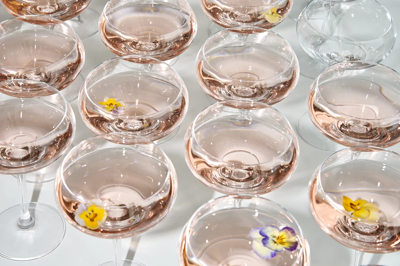 Overhead view of multiple stemmed glasses filled with light pink liquid, some with floating yellow and purple flowers.