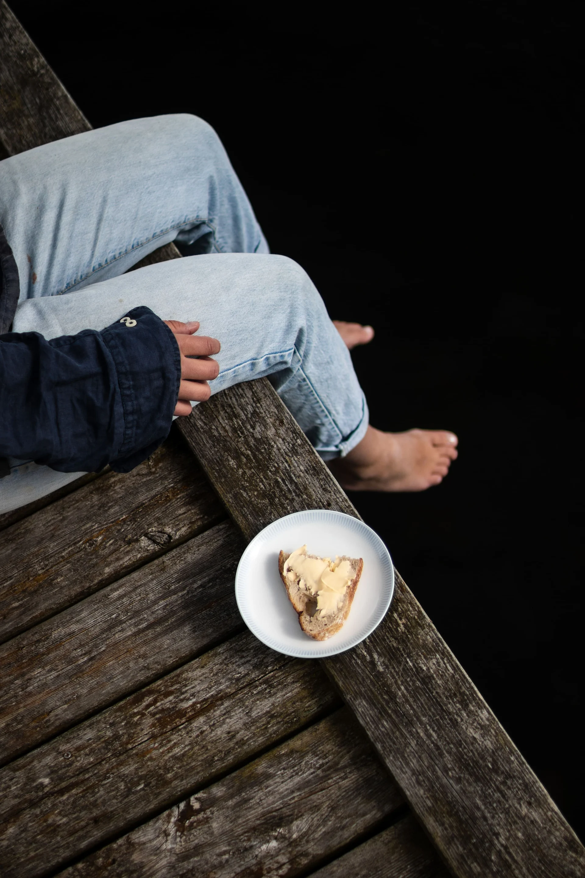 Person sitting on a wooden dock with bare feet next to a plate of bread and butter on a dark background.