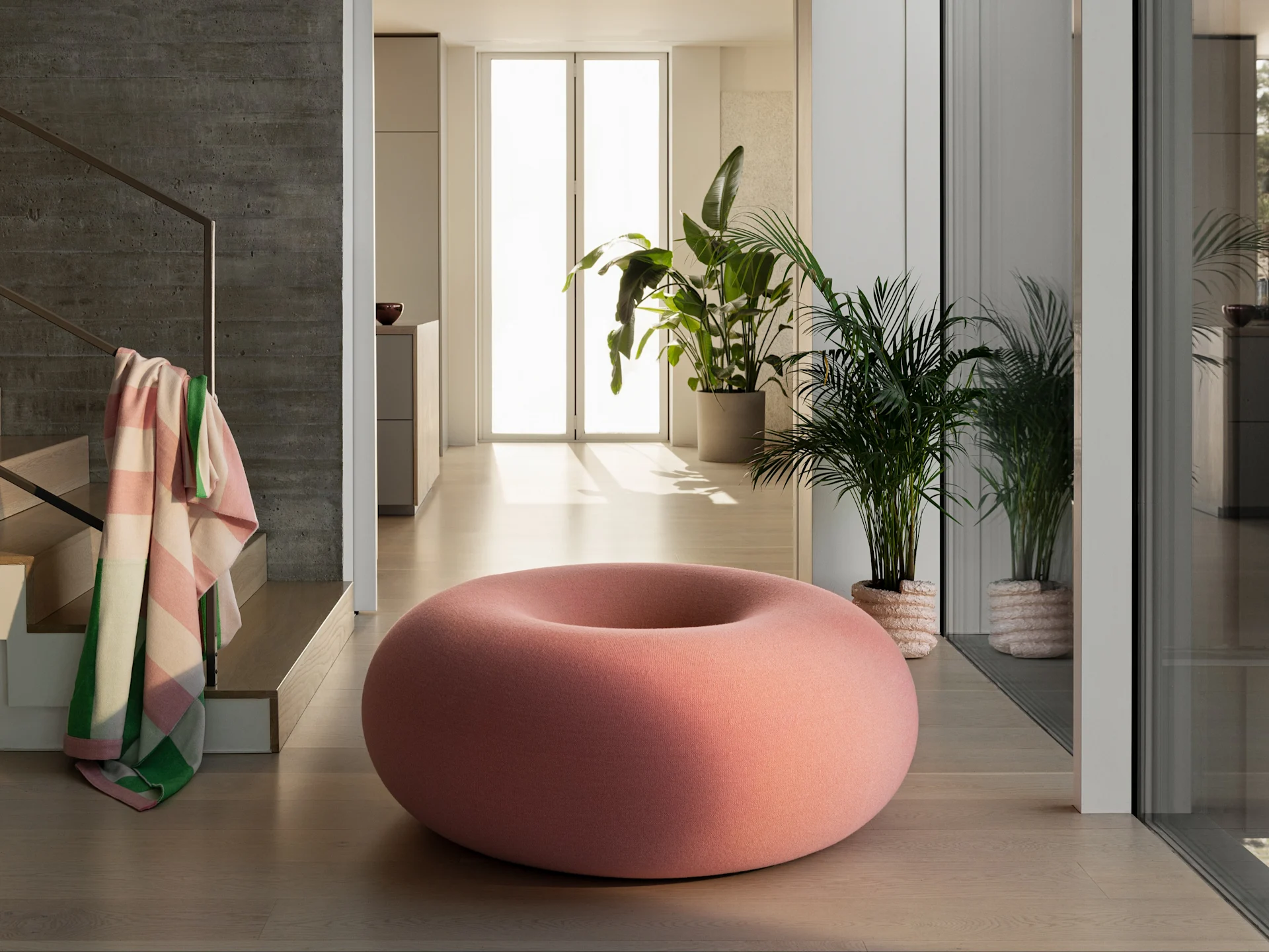 A large, donut-shaped pink pouf on a light wood floor in a modern room with green plants and a concrete wall.