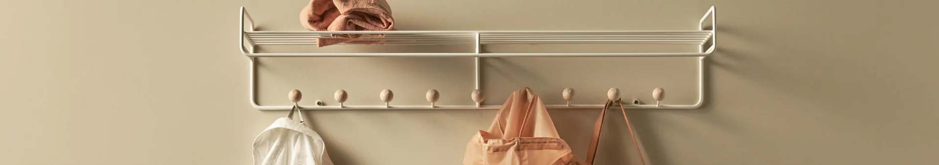 White metal coat rack with wooden pegs, a folded pink towel on top, and a white shirt, pink tote bag, and brown bag hanging.
