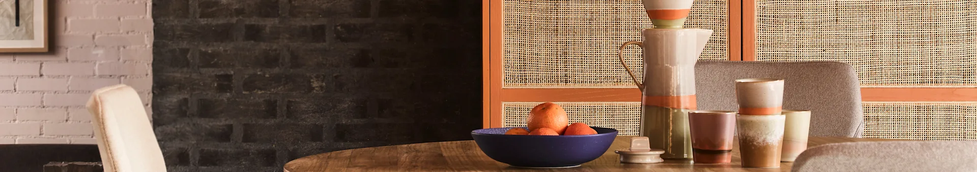 A dining table with a blue bowl of citrus and carrots, surrounded by ceramic pitchers and mugs, against a cane screen and brick wall.