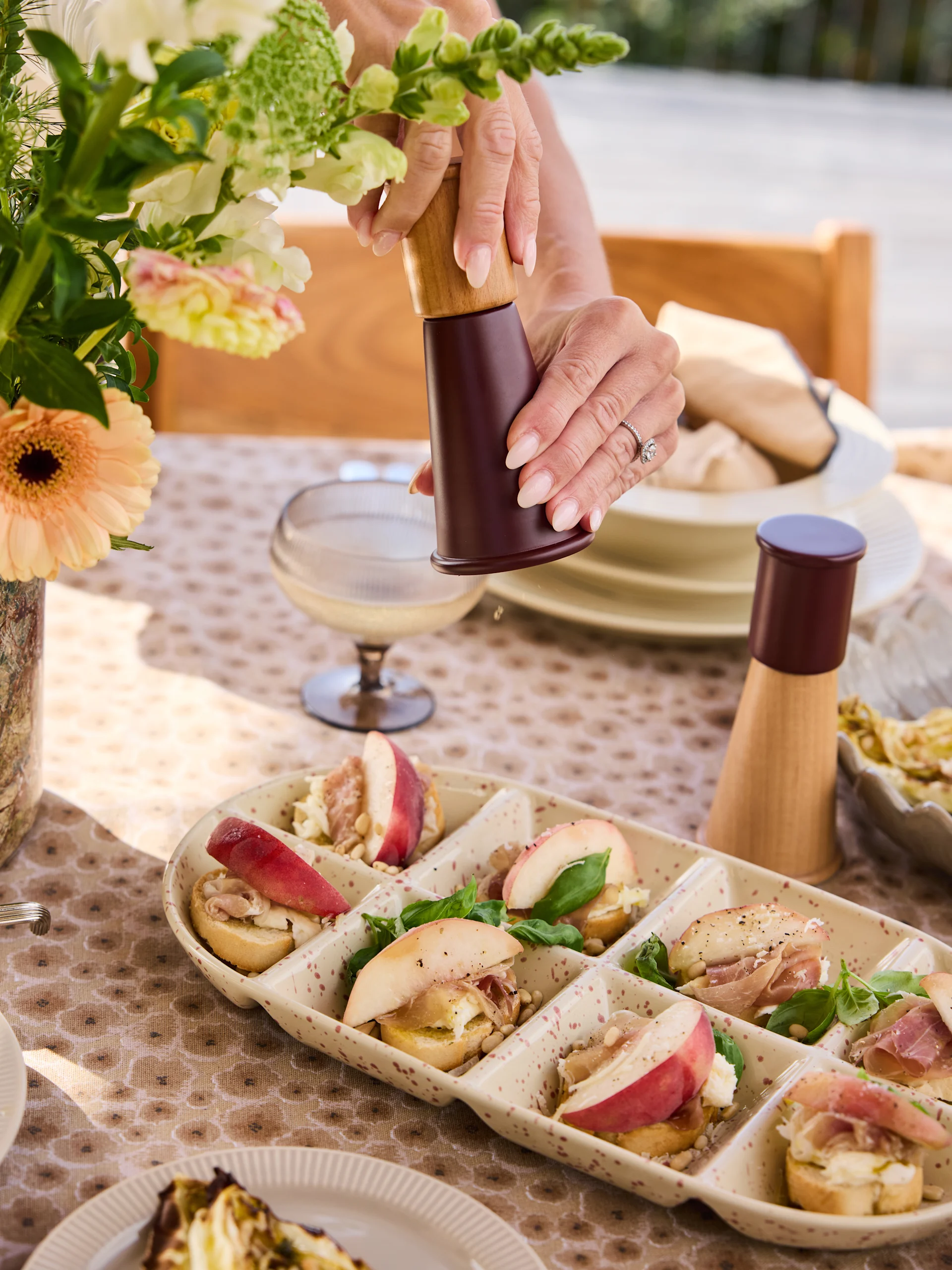 Hands grinding pepper on crostini platter with peaches, prosciutto, cheese, pine nuts, basil.