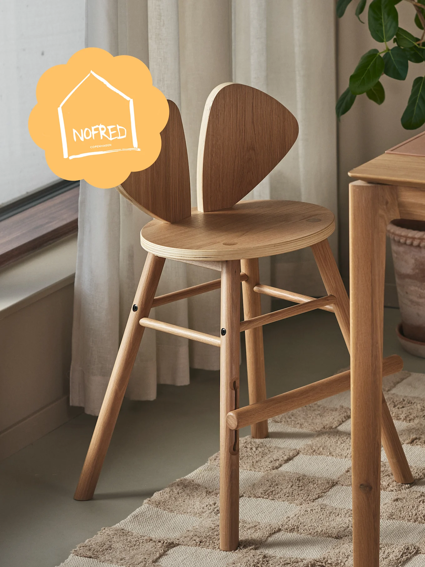 Light wooden high chair with bunny-ear backrest, on a rug, near a wooden table. 'NOFRED COPENHAGEN' logo visible.