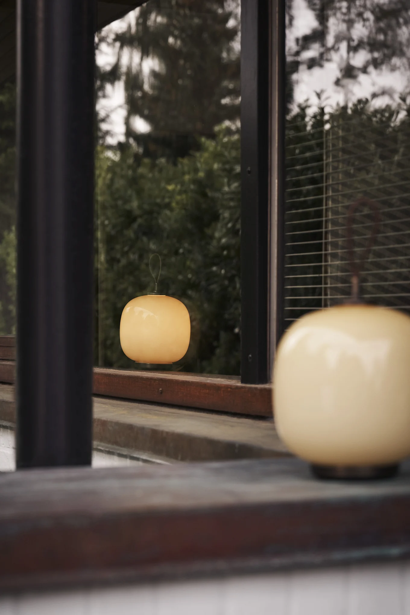 A cream-colored, rounded lamp in the foreground, with another reflected in a window against a green background.
