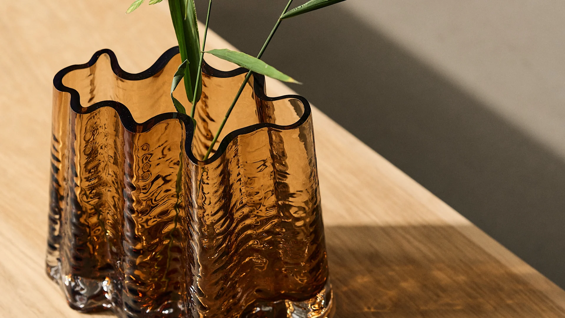 Wavy, amber glass vase with textured surface holding green leaves on a wooden table with a diagonal shadow.