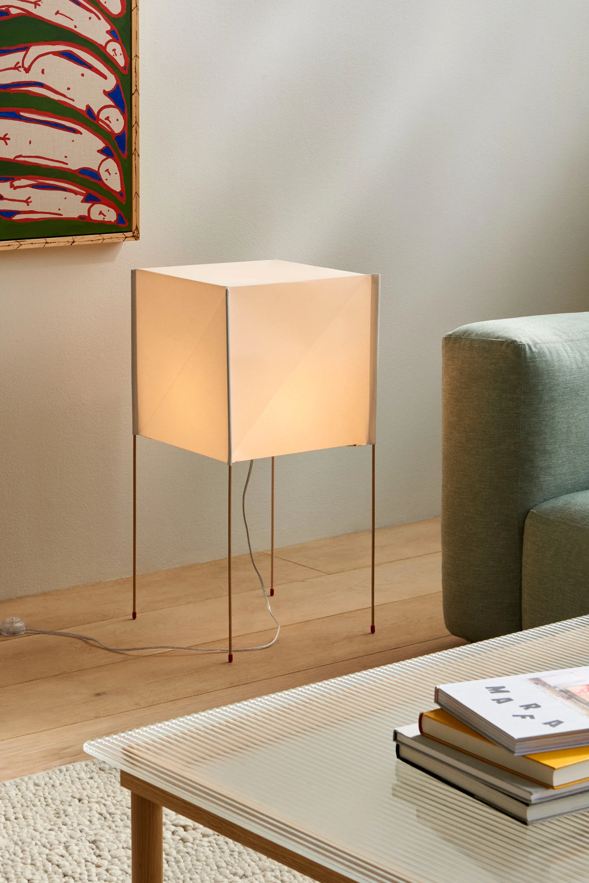 A glowing square floor lamp with thin legs next to a light green sofa and a coffee table with books.