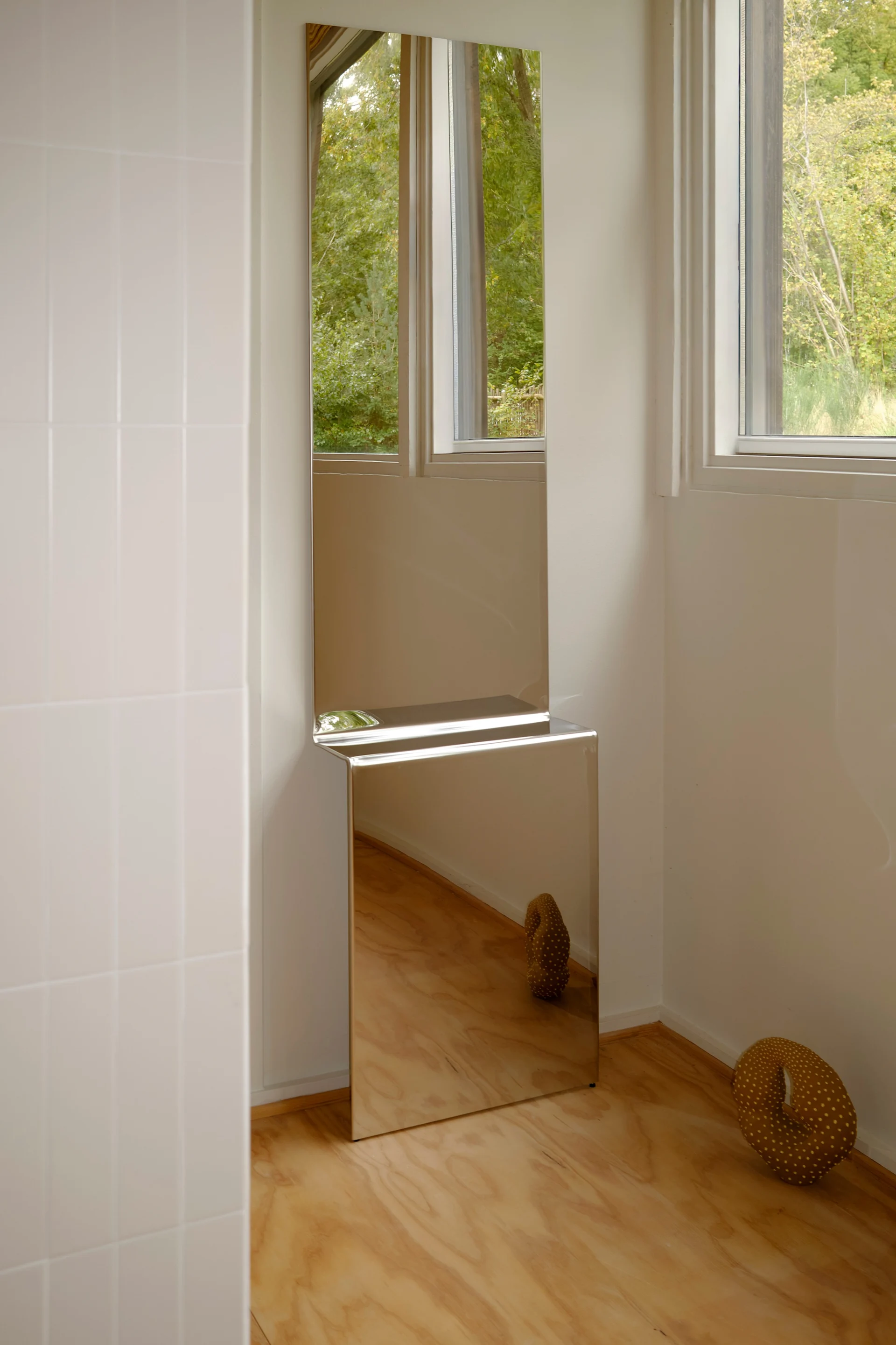 Tall, stacked mirrors in a modern room. The top reflects a window and trees; the bottom reflects a wooden floor and a brown dotted sculpture.