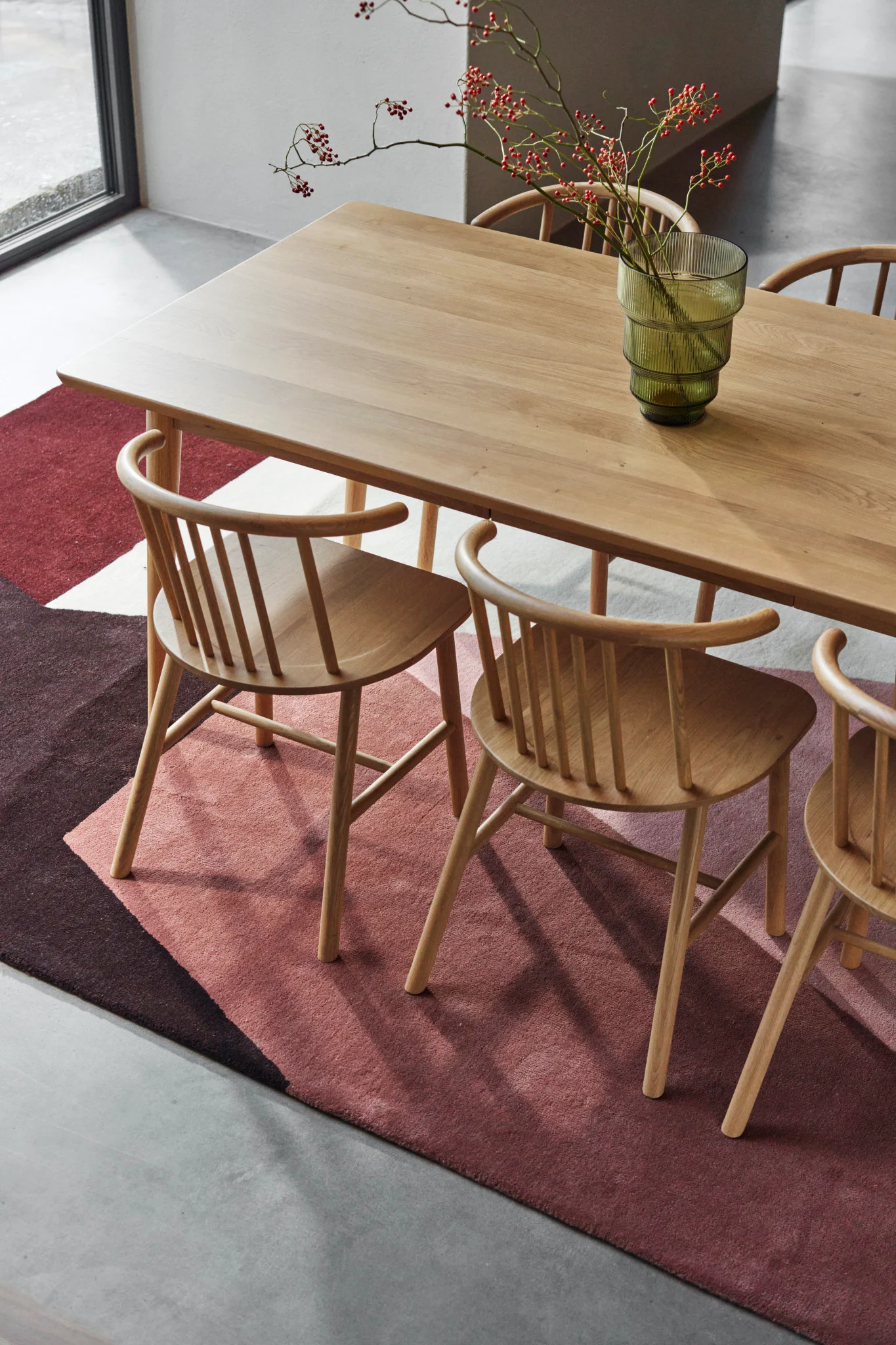 A modern dining room with a light wood table, matching chairs, a geometric red and pink rug, and a vase of red berries.