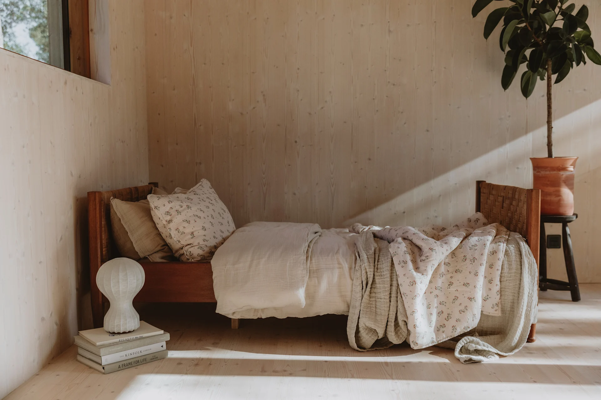 Cozy bedroom with wood walls, a wooden bed with cream and floral bedding, books with a lamp, and a potted plant.