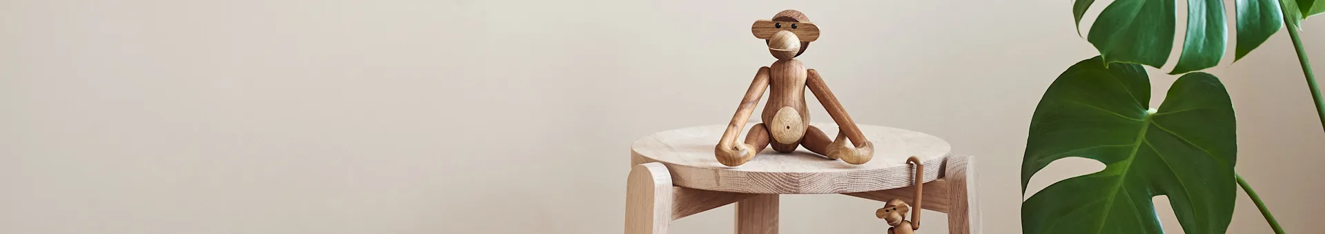 A large wooden monkey figurine sits on a light wooden stool, with a smaller wooden monkey hanging below it, next to a green plant.