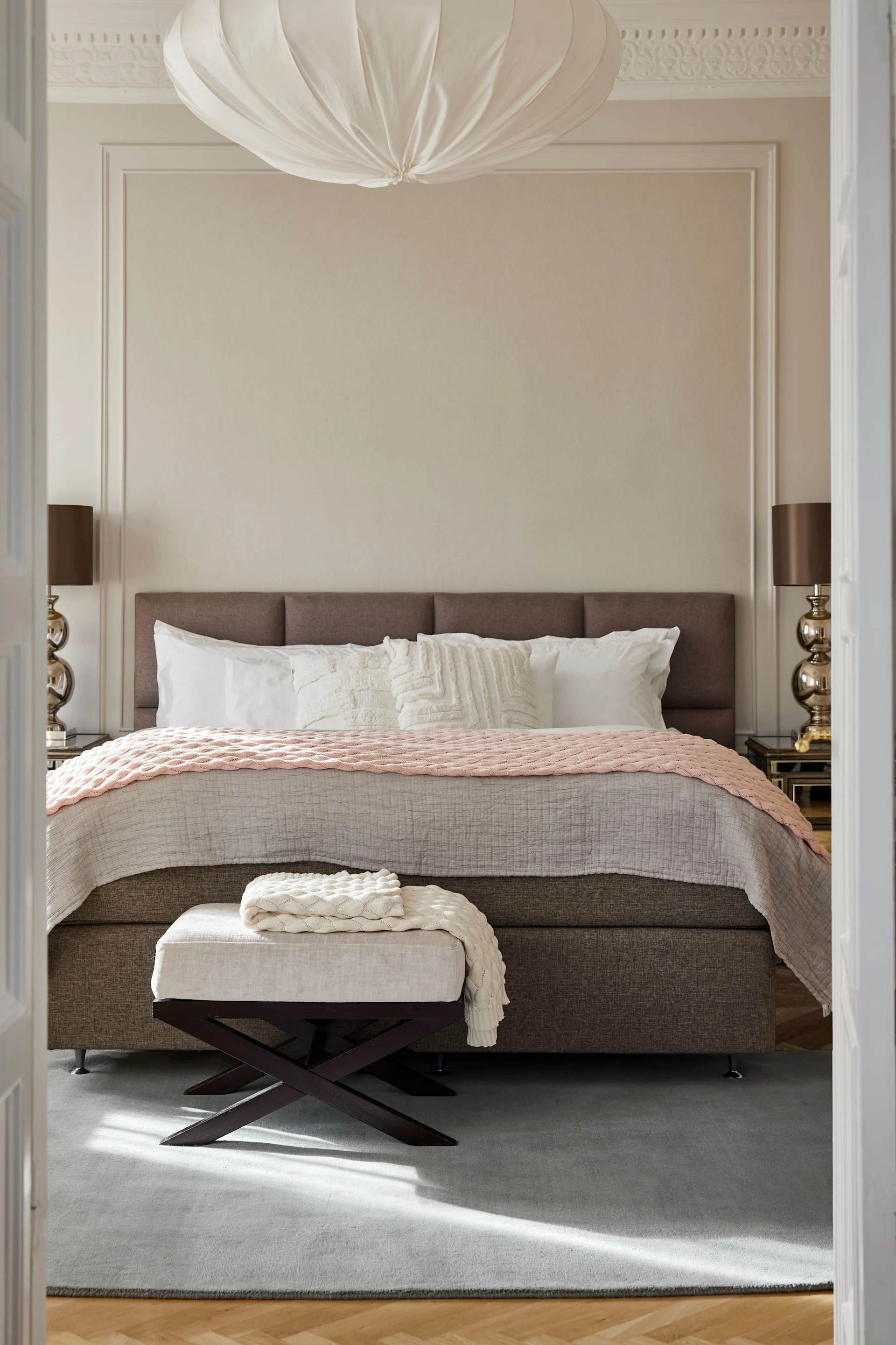 Elegant bedroom with a brown bed, pink and grey bedding, a white bench, and two lamps, viewed through an open door.