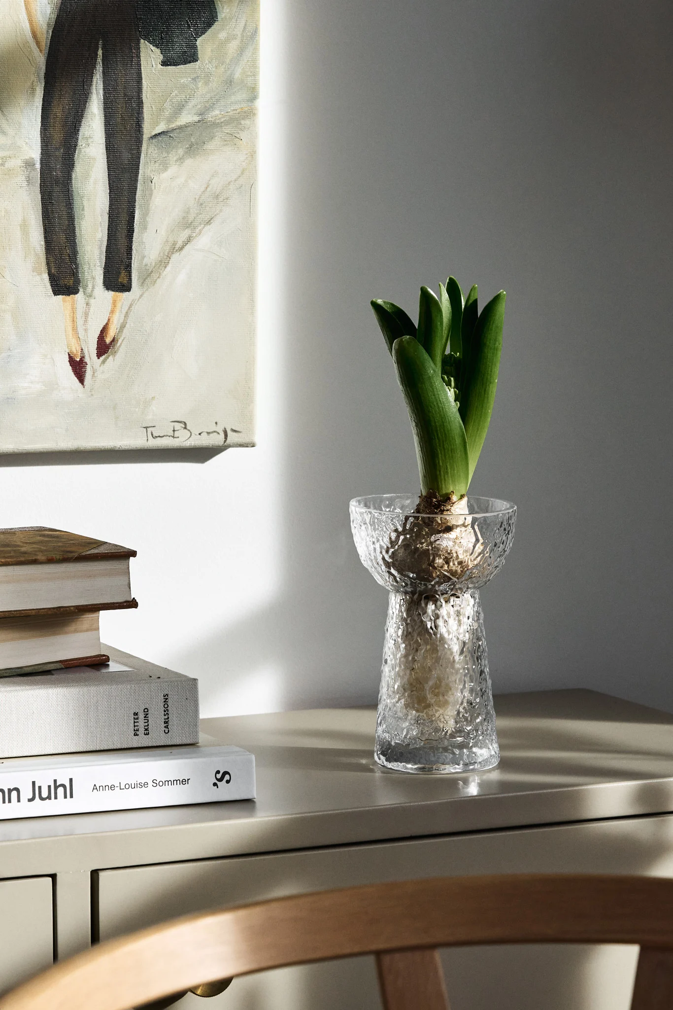 Green hyacinth plant in a textured glass vase on a light cabinet, next to a painting of legs and books.