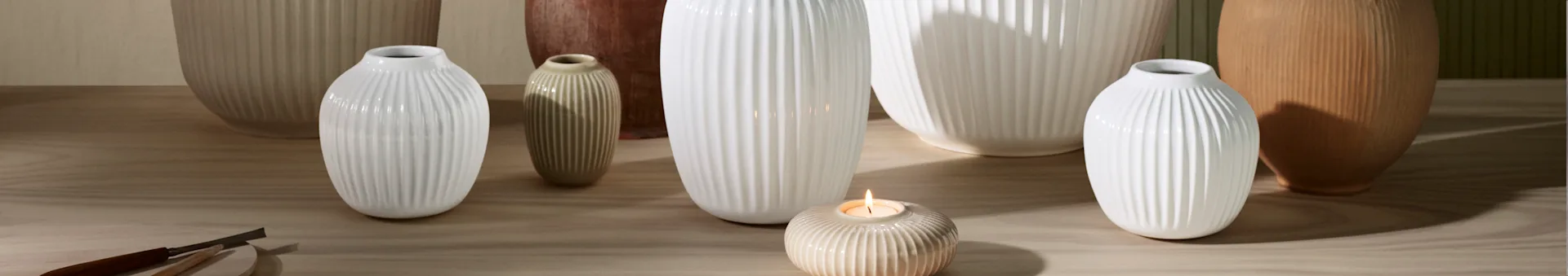 A collection of ribbed white and cream vases and a lit candle in a ribbed candle holder on a wooden surface.