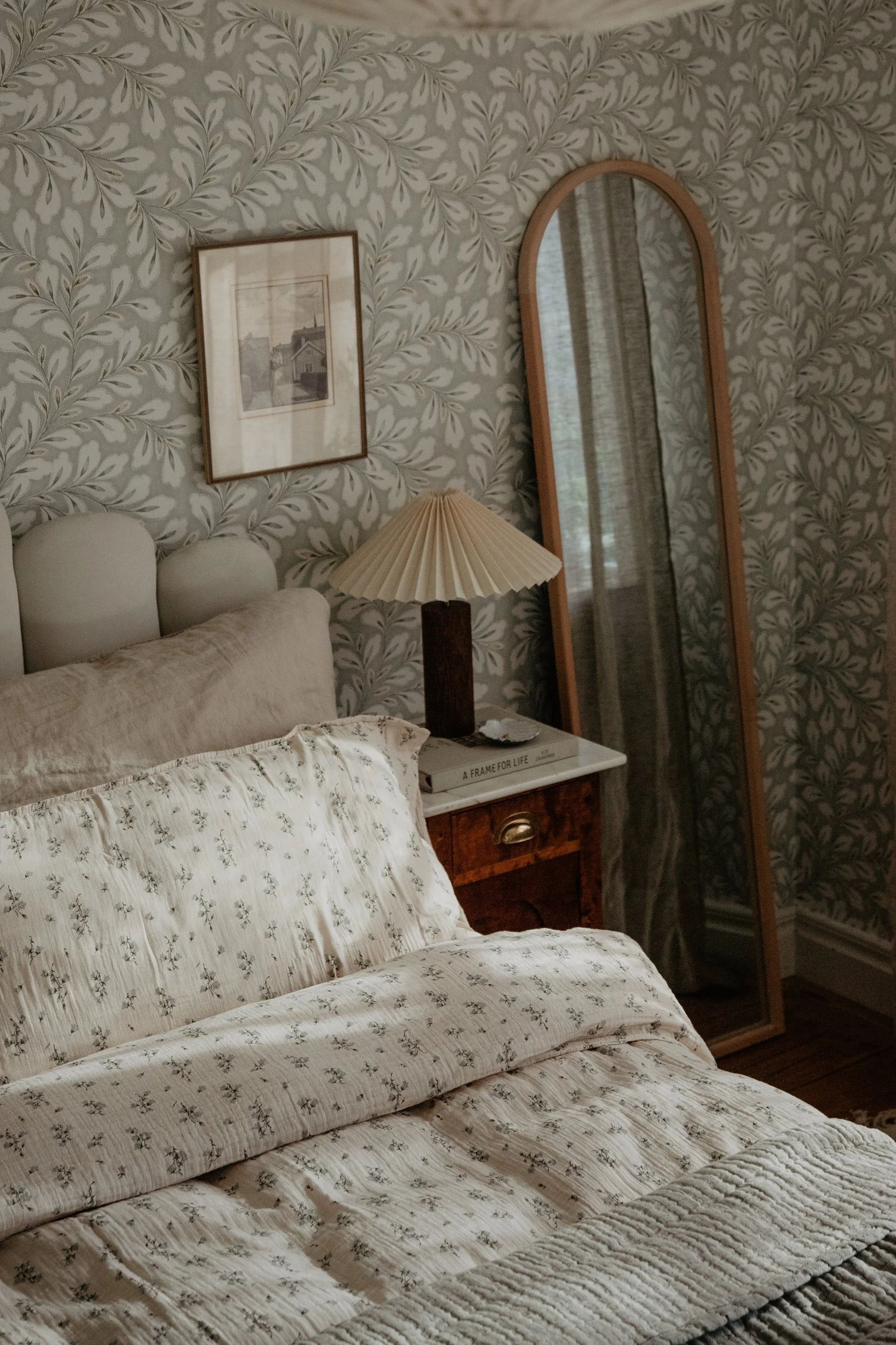 A bedroom with cream patterned bedding, a pleated lamp on a wooden nightstand, and a tall mirror, against leafy wallpaper.