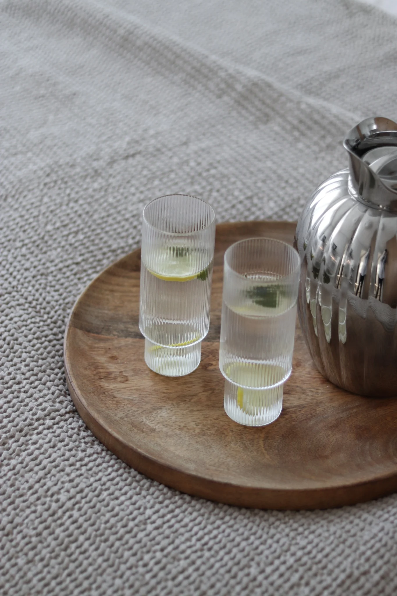 Two ribbed glasses of water with lemon and mint, and a silver fluted pitcher on a round wooden tray, on a beige woven blanket.