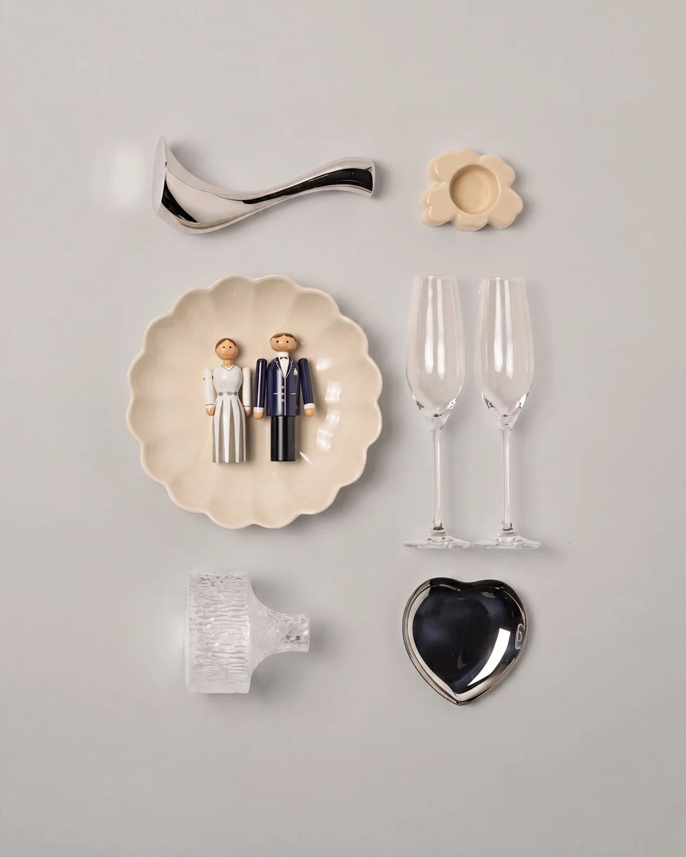 Wedding flat lay with wooden bride and groom figurines on a scalloped plate, two champagne flutes, a silver heart dish, and assorted decor.