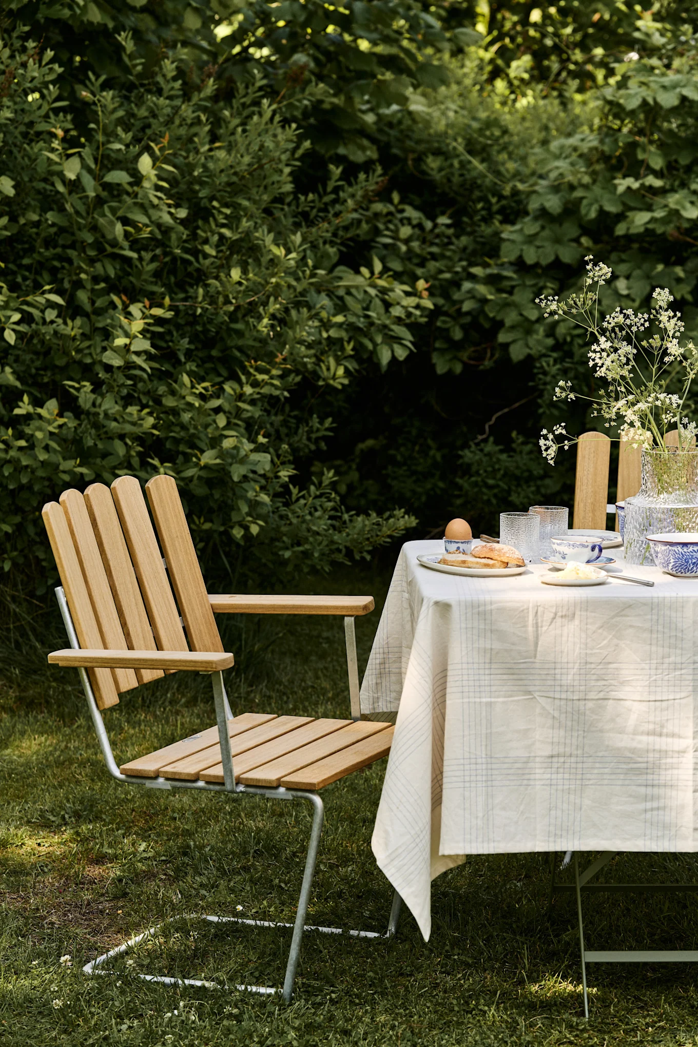 An outdoor breakfast scene with a light wood slat and metal chair next to a table set with plaid tablecloth, dishes, and a vase of white flowers.