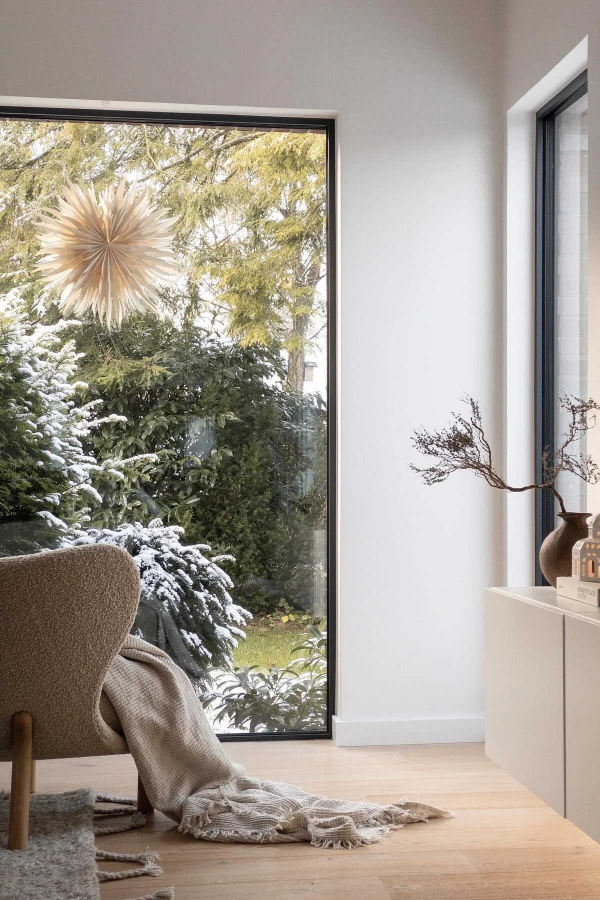 Cozy room with a large window overlooking a snowy garden. An armchair with a throw blanket sits on a wooden floor, near a console table with dried branches.