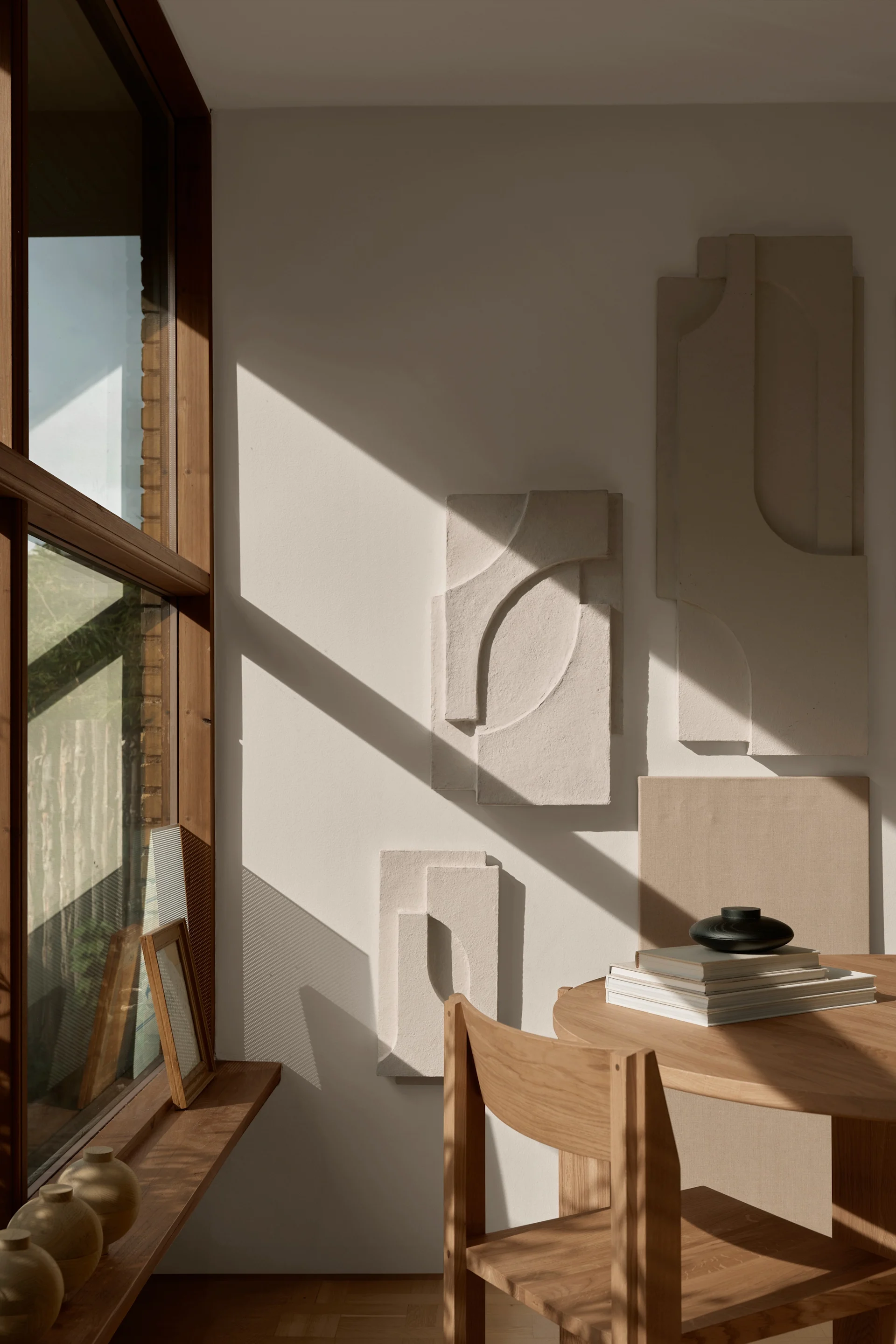 Sunny room with abstract wall art, a wooden table, and a black vase on books, next to a large window.