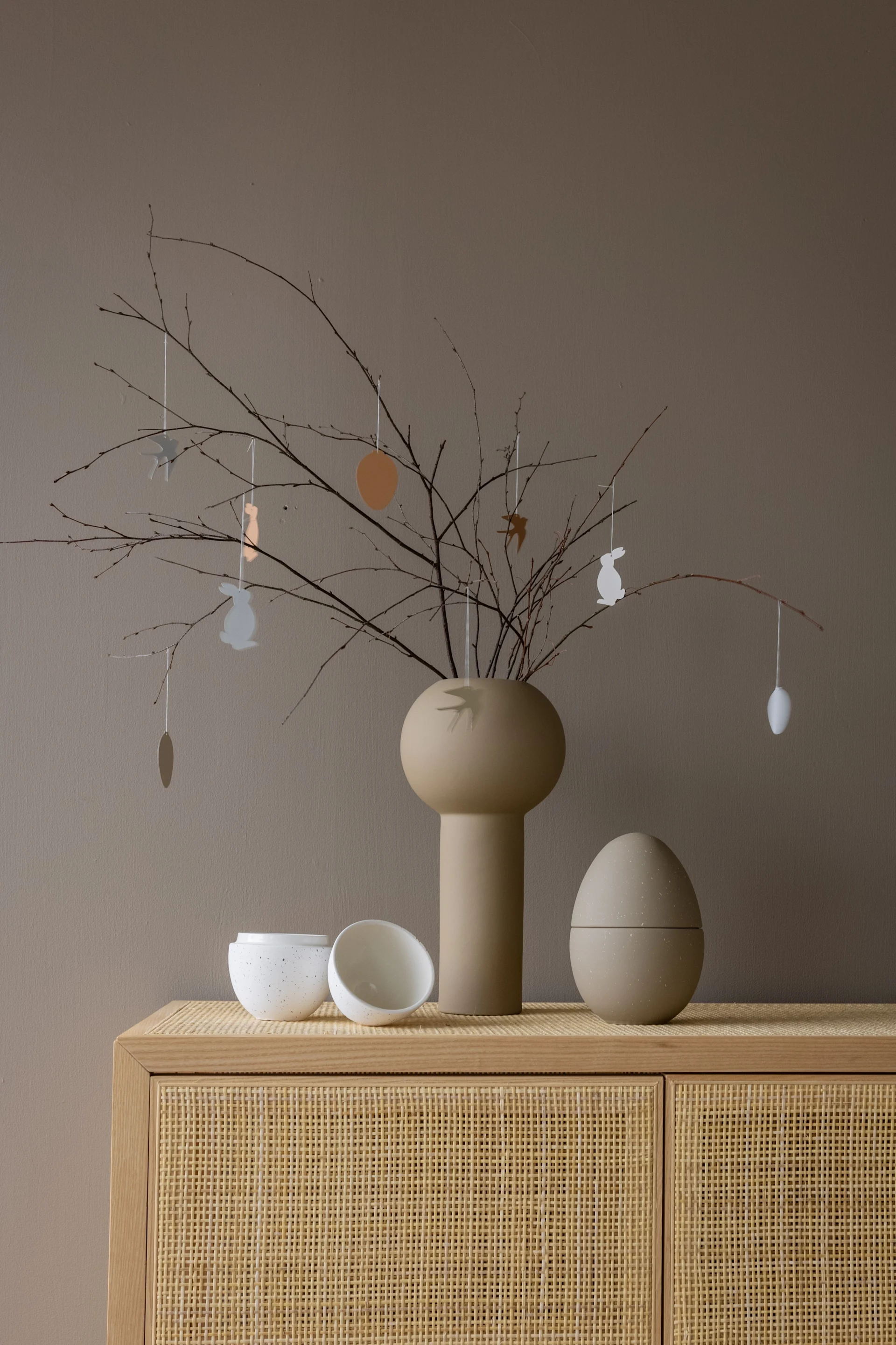 Beige vase with Easter branches, hanging bunnies, eggs, birds. White bowls, large beige egg on rattan cabinet.