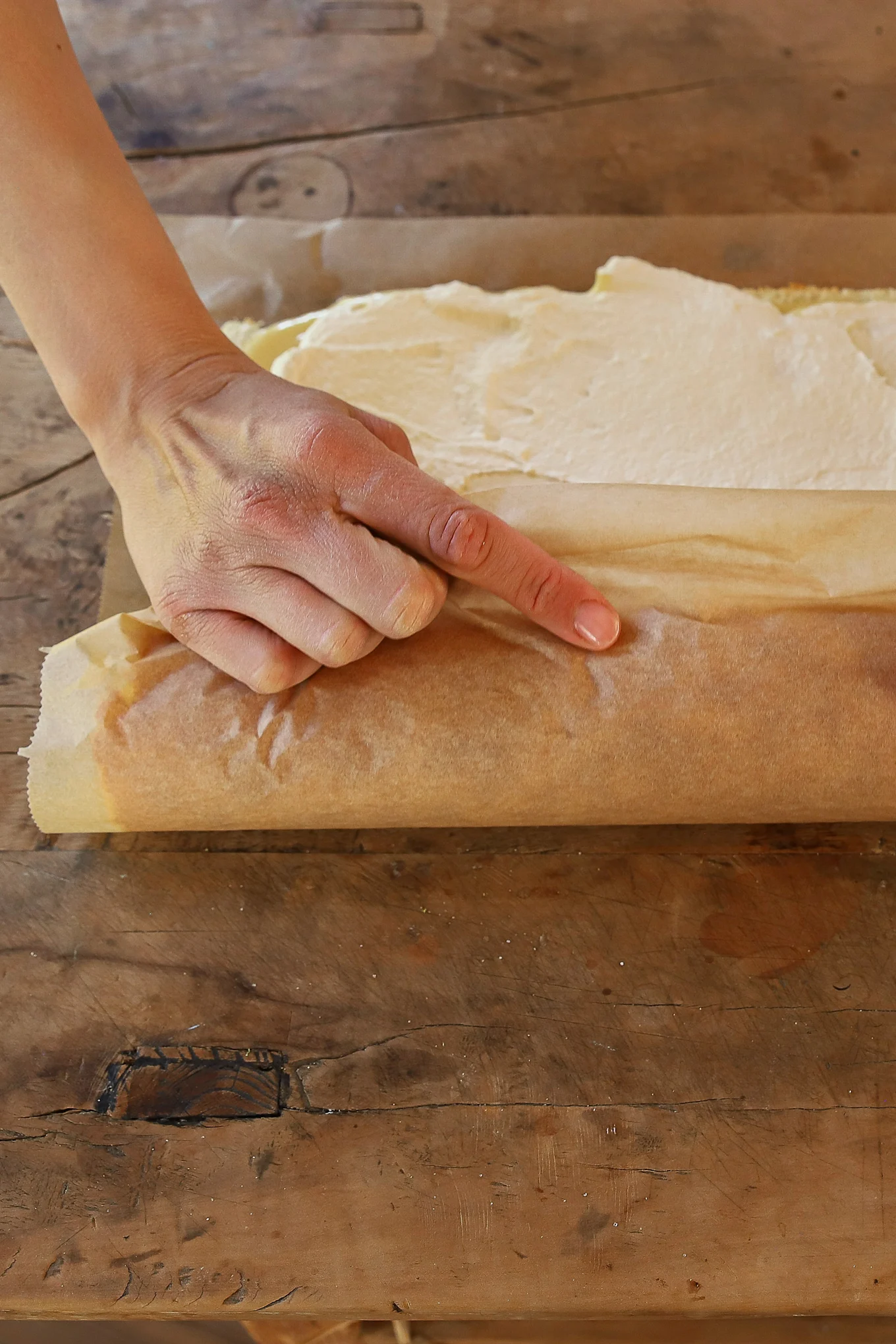 A hand rolling a creamy dessert on parchment paper on a wooden table.