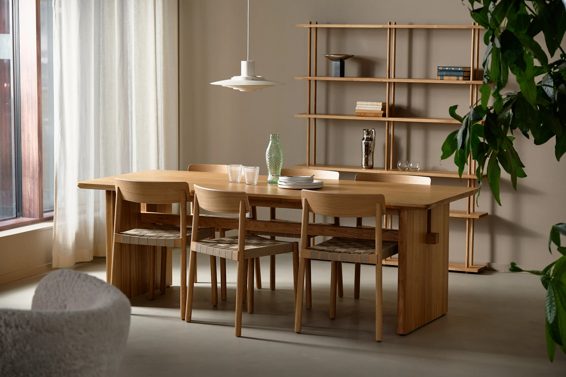 A modern dining room with a long wooden table, six chairs, a tiered pendant lamp, and a wooden open shelf with decor.