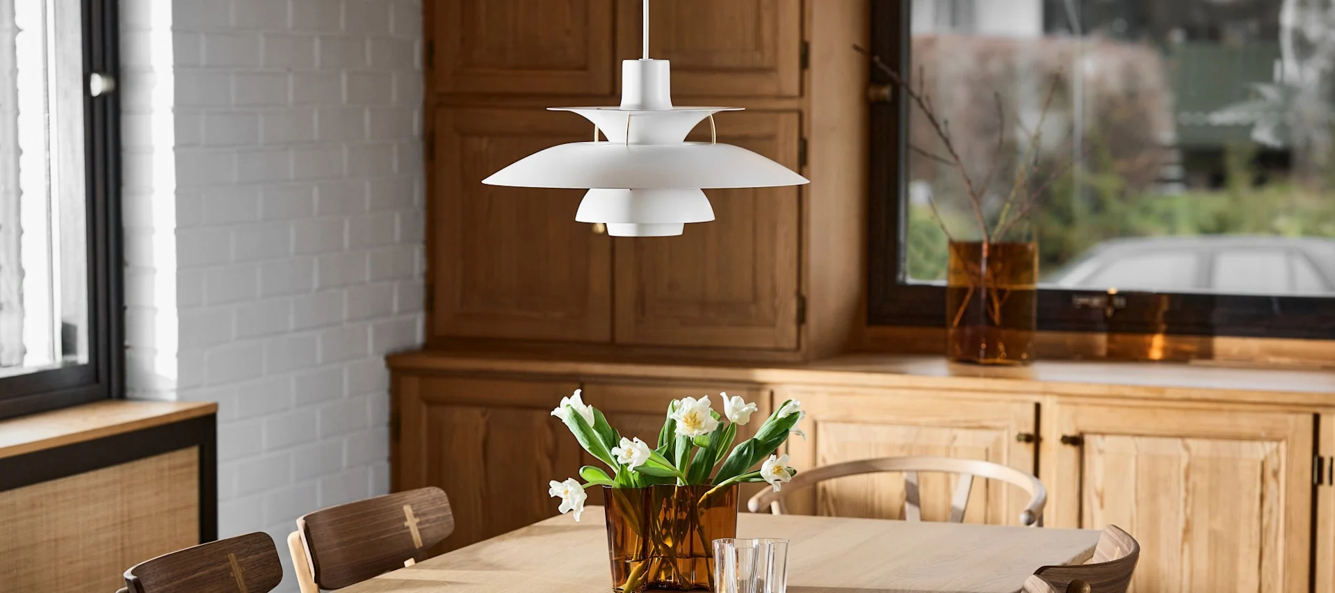 A white, multi-tiered pendant lamp hangs over a wooden table with a vase of white tulips, in a room with wooden cabinets.