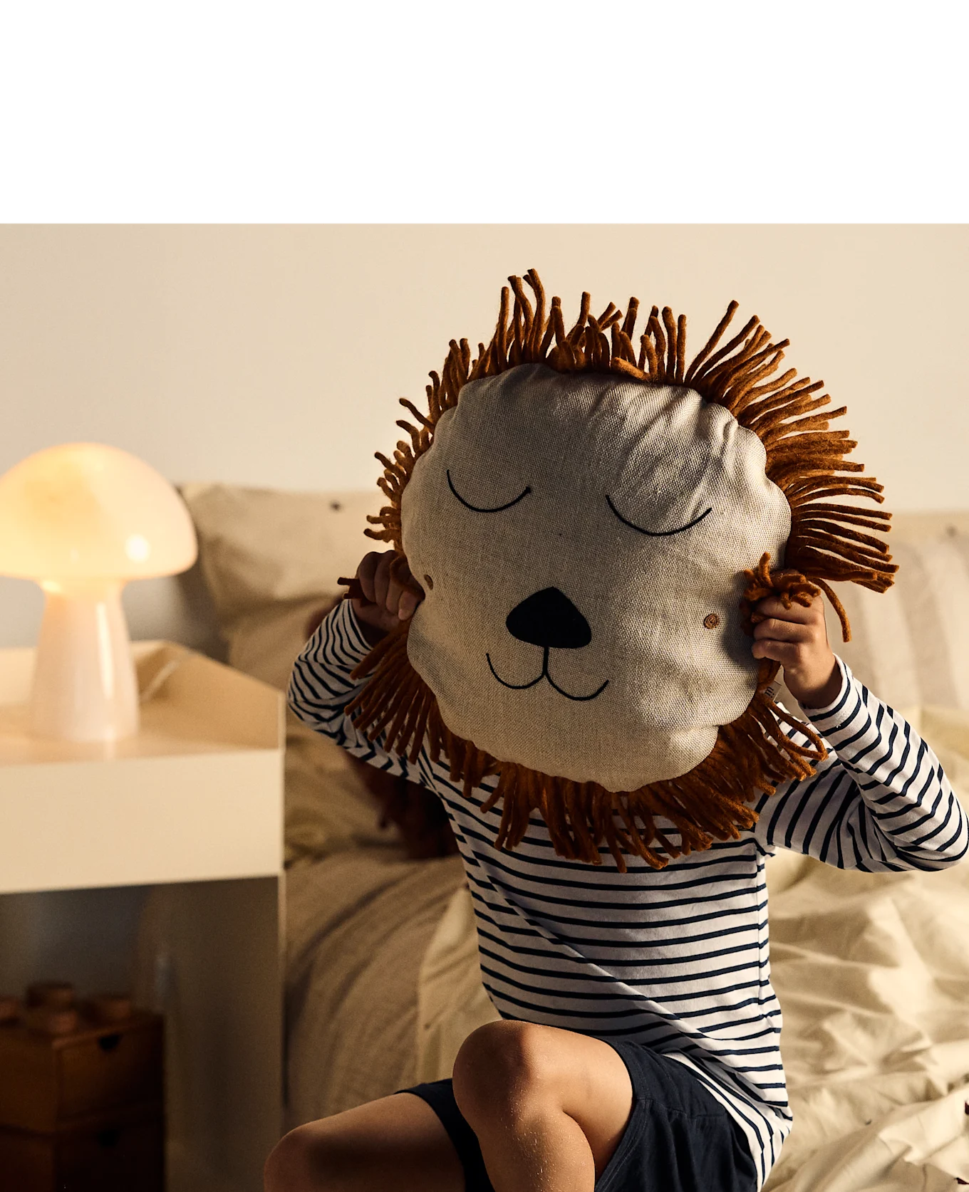 Child holding a lion-face pillow over their face while sitting on a bed with a glowing mushroom lamp nearby.