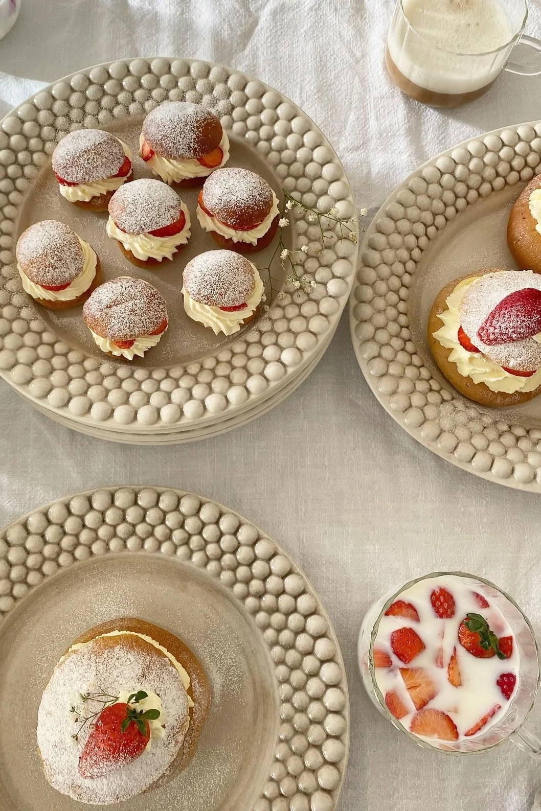 Cream buns with whipped cream, strawberries, and powdered sugar on textured plates, with drinks.