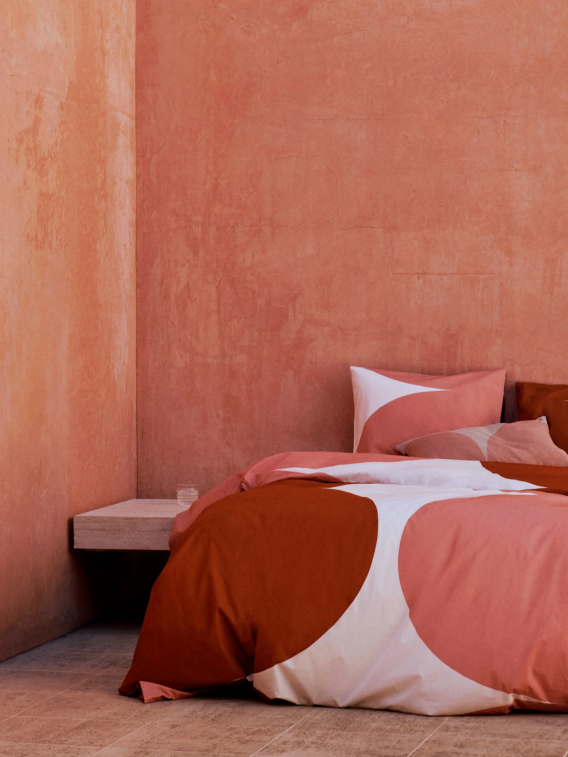 Bed with terracotta, white, and coral abstract pattern bedding in a room with textured reddish-orange walls and tiled floor.