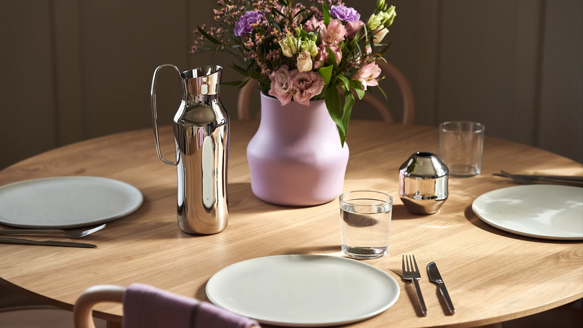 A round wooden table set for two with light plates, silverware, a metallic pitcher, a purple vase with flowers, and a glass of water.