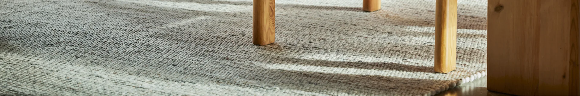 Close-up of a textured, light-colored rug with two light brown wooden furniture legs casting shadows on it.