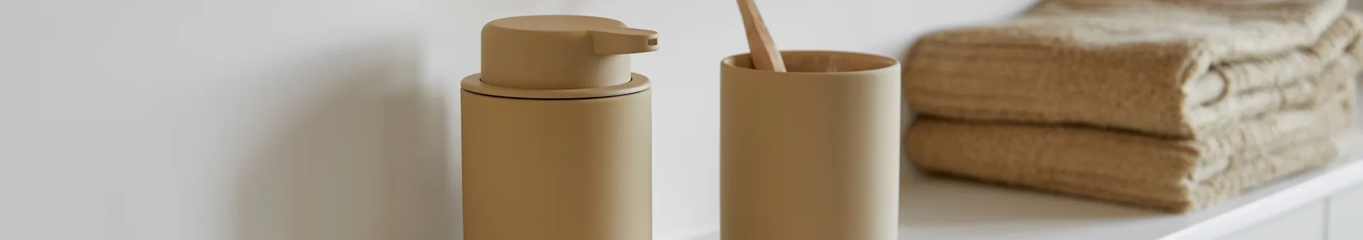 Beige soap dispenser and toothbrush holder with a wooden toothbrush, beside stacked beige towels on a white surface.