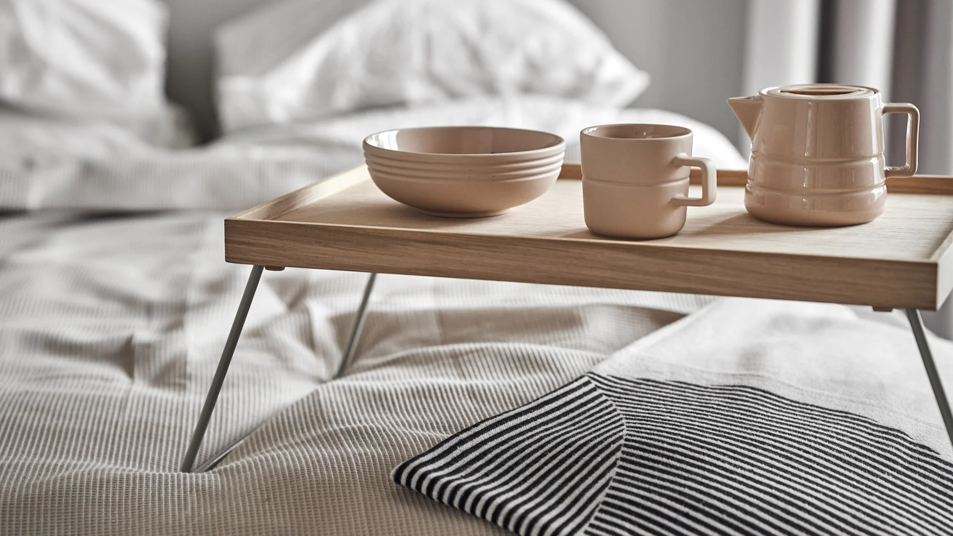 A light brown bowl, mug, and teapot on a wooden breakfast tray on a bed with white bedding and a striped textile.