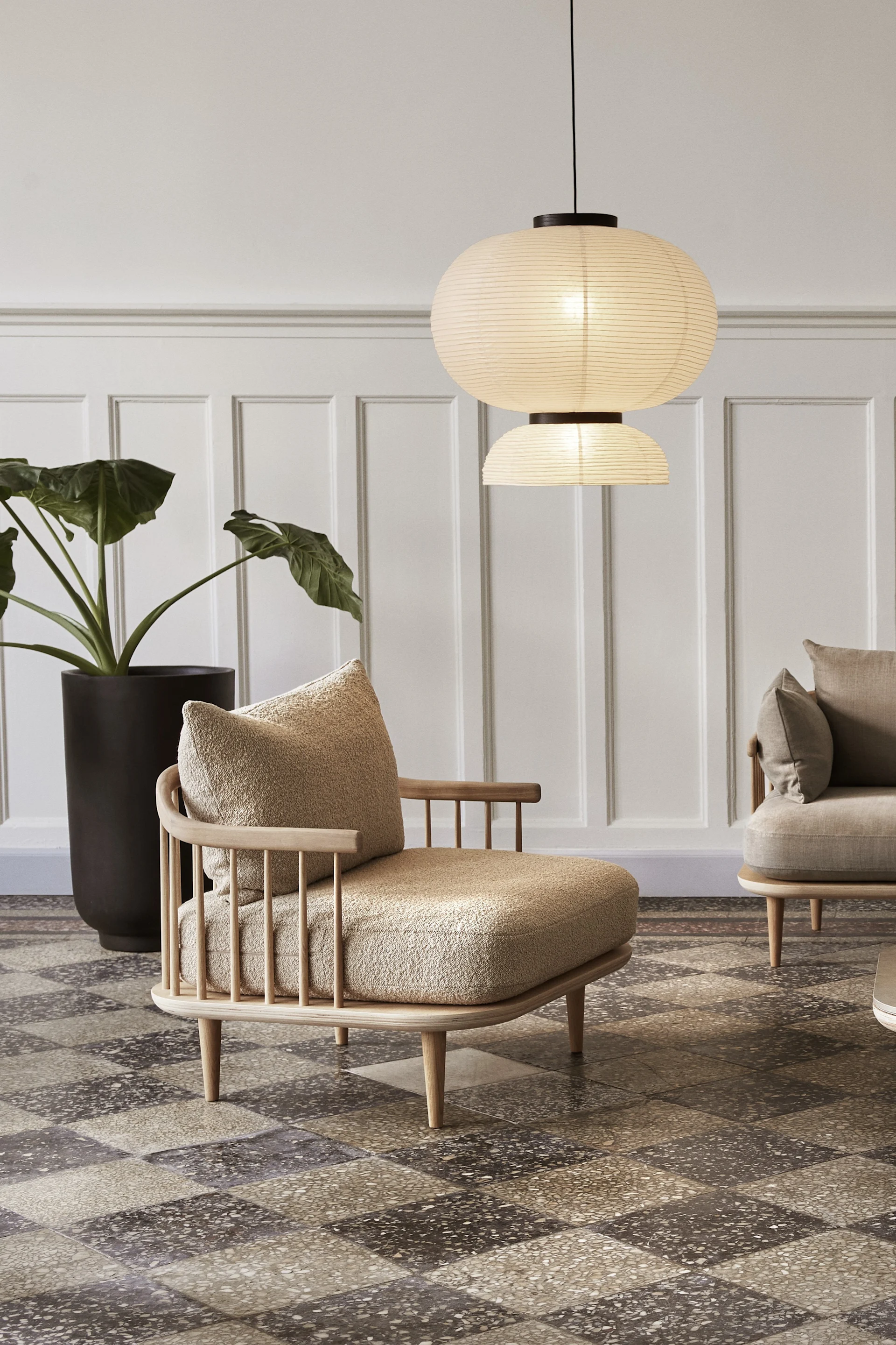 A modern living room featuring a light wood armchair with textured beige cushions, two layered paper lantern pendant lights, and a large potted plant.