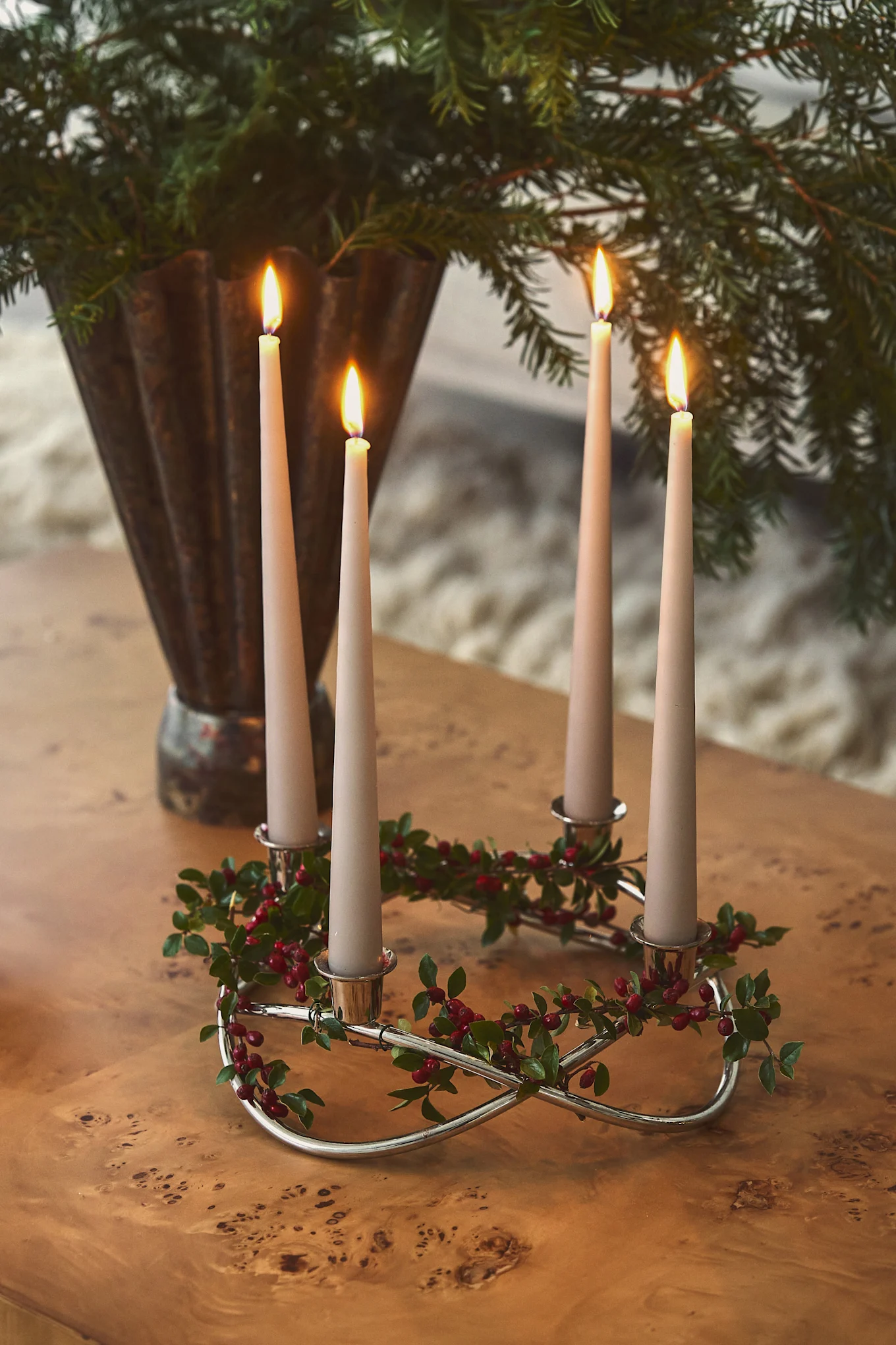 Four lit candles in a silver Advent wreath with green leaves and red berries, on a wooden table. Evergreen in background.