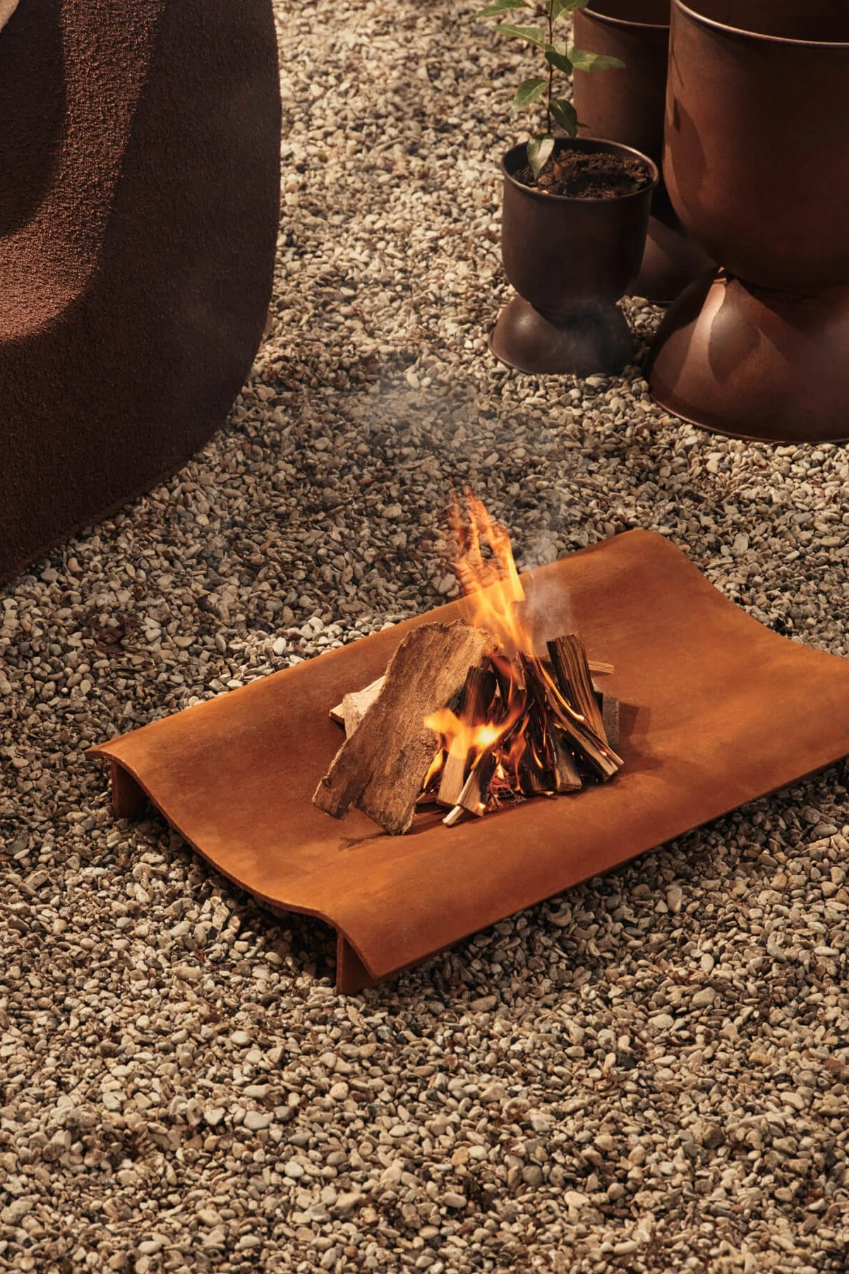 A small campfire burns on a curved, rusty orange metal tray set on a gravel surface.