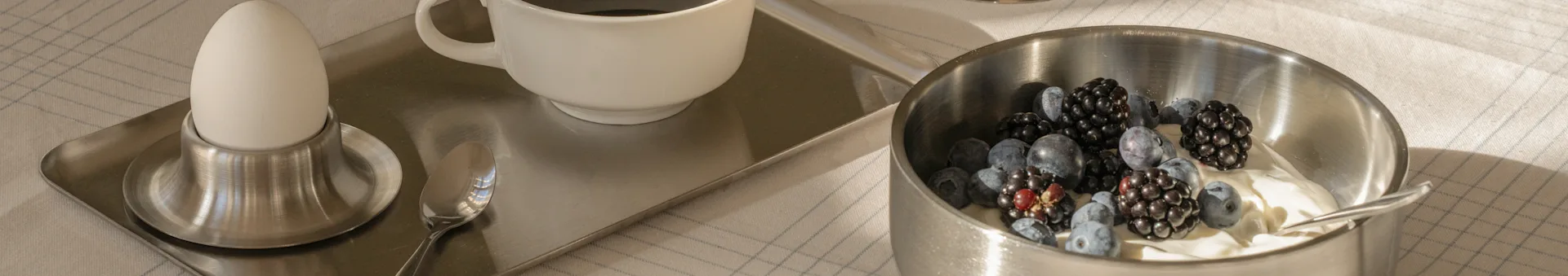 Breakfast tray with a boiled egg, coffee, and a bowl of yogurt topped with blueberries and blackberries on a checkered tablecloth.