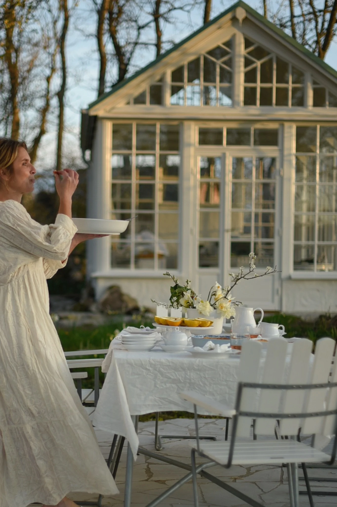 Woman holds a platter at an outdoor table set with white dishes, flowers, and lemons, with a greenhouse in the background.